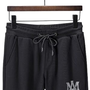 May include: Black sweatpants with a drawstring waist and side pockets. The pants are hanging on a wooden hanger. A silver-colored logo is visible on the lower leg.