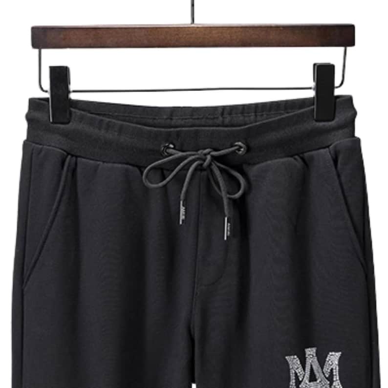 May include: Black sweatpants with a drawstring waist and side pockets. The pants are hanging on a wooden hanger. A silver-colored logo is visible on the lower leg.
