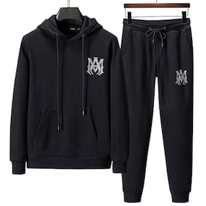 May include: A black hoodie and matching sweatpants set. The hoodie has a front pocket and a drawstring hood. Both pieces feature a silver-colored emblem on the front. The sweatpants have a drawstring waist.