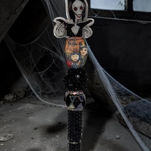 May include: A decorative pen featuring a character topper with black hair and red button eyes, followed by an oval charm depicting a group of illustrated figures, a black textured bead, a faceted crystal bead, and a black rhinestone-encrusted barrel.