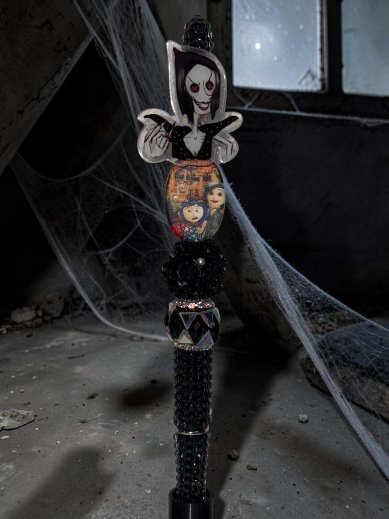 May include: A decorative pen featuring a character topper with black hair and red button eyes, followed by an oval charm depicting a group of illustrated figures, a black textured bead, a faceted crystal bead, and a black rhinestone-encrusted barrel.