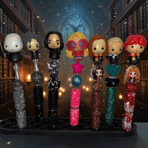 May include: A set of seven decorative pens featuring character toppers, including figures with various hair colors and outfits. Each pen barrel is covered in sparkling rhinestones in colors like silver, black, pink, brown, green, and red, arranged on a black stand.