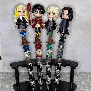 May include: Four decorative pens featuring characters from a popular fantasy series. Each pen has a character figurine, a wizard hat, and a beaded design in various colors. The pen barrels are black and silver with a crystal-like pattern.