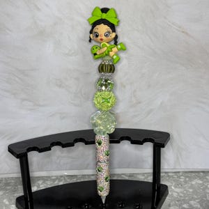K&ouml;nnte beinhalten: Dekorativer Stift mit einer Cartoon-Figur eines M&auml;dchens mit gr&uuml;ner Schleife und Kleid, das einen Frosch h&auml;lt. Der Stift hat einen silbernen Schaft, der mit gr&uuml;nen Edelsteinen und einer Reihe von gr&uuml;nen und klaren Perlen verziert ist. Der Stift wird auf einem schwarzen St&auml;nder ausgestellt.