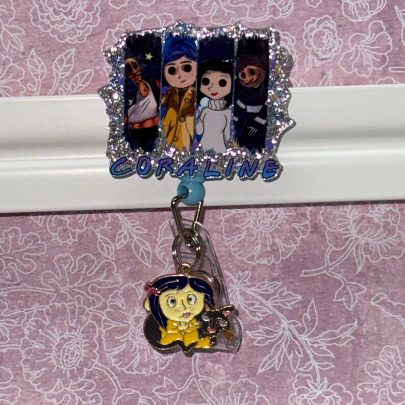 May include: A badge reel featuring a rectangular acrylic topper with a glittery border and four character illustrations from Coraline, with the text CORALINE printed below. A metal clip attaches a small enamel charm of the character Coraline and a black cat.