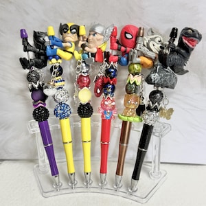 May include: A collection of decorative pens featuring superhero character toppers. The pens have colorful barrels and beaded accents. Characters include Black Panther, Wolverine, Thor, Spider-Man, Rocket Raccoon, and Godzilla.