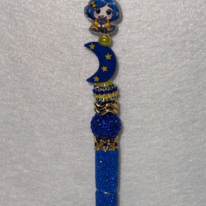 May include: A decorative pen featuring a blue rhinestone-covered barrel and a series of stacked beads. The topper includes a blue crescent moon with yellow stars, a small yellow bead, and a character charm with blue hair holding a black cat.