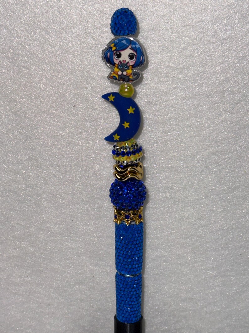 May include: A decorative pen featuring a blue rhinestone-covered barrel and a series of stacked beads. The topper includes a blue crescent moon with yellow stars, a small yellow bead, and a character charm with blue hair holding a black cat.