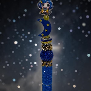 May include: A decorative pen topper featuring a blue crescent moon with yellow stars, a cartoon character with blue hair holding a black cat, and a series of stacked beads in shades of blue, gold, and silver with a textured, sparkling finish.
