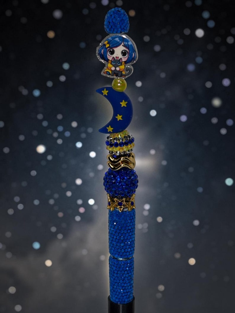 May include: A decorative pen topper featuring a blue crescent moon with yellow stars, a cartoon character with blue hair holding a black cat, and a series of stacked beads in shades of blue, gold, and silver with a textured, sparkling finish.