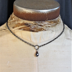 May include: A black chain necklace with a teardrop-shaped pendant. The pendant is a dark red color with a faceted surface.