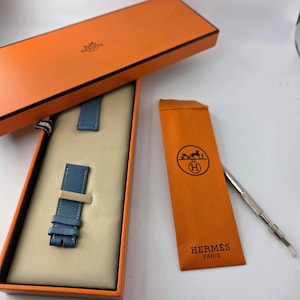 Könnte beinhalten: Ein blaues Leder-Uhrenarmband in einer orangefarbenen Schachtel mit dem Markennamen "Hermès Paris" auf der Schachtel und dem Beutel. Ein silbernes Werkzeug ist ebenfalls enthalten. Das Uhrenarmband wird auf einem beigen Innenraum präsentiert.