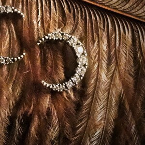 Op de afbeelding: Een paar zilveren halvemaanvormige oorbellen met een rij heldere, glinsterende stenen rond de rand.