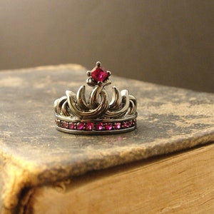 Peut inclure: Une couronne en argent avec un joyau rose sur le dessus et une rang&eacute;e de joyaux roses autour de la base. La couronne est pos&eacute;e sur un vieux livre brun.