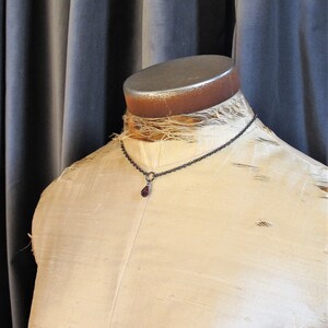 May include: A silver chain necklace with a dark red teardrop pendant. The necklace is displayed on a white fabric-covered mannequin.