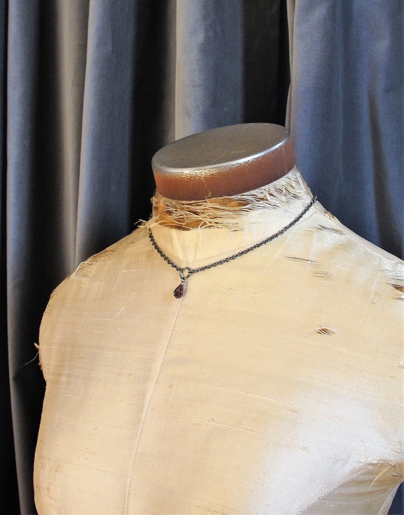 May include: A silver chain necklace with a dark red teardrop pendant. The necklace is displayed on a white fabric-covered mannequin.