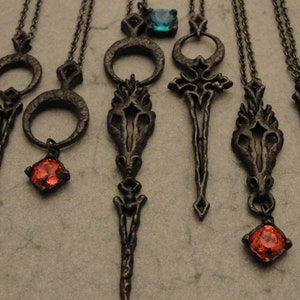 May include: Seven black metal pendants with intricate designs, some featuring a red gemstone and one with a blue gemstone. The pendants are on chains and appear to be jewelry.