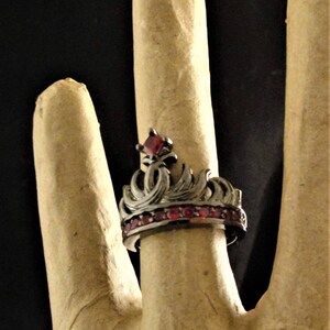 Peut inclure: Une bague en forme de couronne noire avec des pierres pr&eacute;cieuses rouges. La couronne est orn&eacute;e de motifs complexes et la bande est sertie de petites pierres rouges.