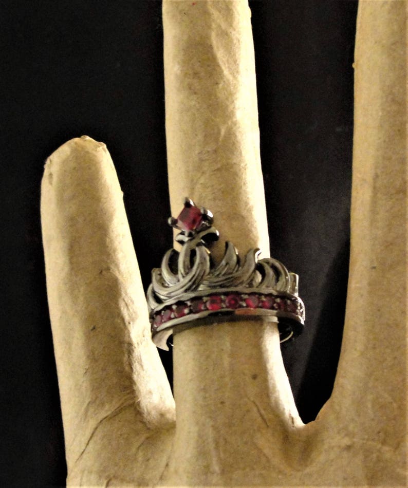 Peut inclure: Une bague en forme de couronne noire avec des pierres pr&eacute;cieuses rouges. La couronne est orn&eacute;e de motifs complexes et la bande est sertie de petites pierres rouges.
