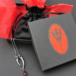 Peut inclure: Un collier en argent avec un pendentif en forme de deux cornes avec une pierre pr&eacute;cieuse rouge &agrave; l'extr&eacute;mit&eacute;. Le collier est dans une bo&icirc;te noire avec un papier de soie rouge et un logo sur le couvercle.