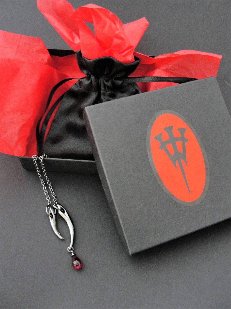 Peut inclure: Un collier en argent avec un pendentif en forme de deux cornes avec une pierre pr&eacute;cieuse rouge &agrave; l'extr&eacute;mit&eacute;. Le collier est dans une bo&icirc;te noire avec un papier de soie rouge et un logo sur le couvercle.