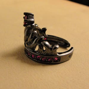 Peut inclure: Une bague couronne noire avec une seule pierre rouge sur le dessus et une rang&eacute;e de petites pierres rouges autour de la bande.