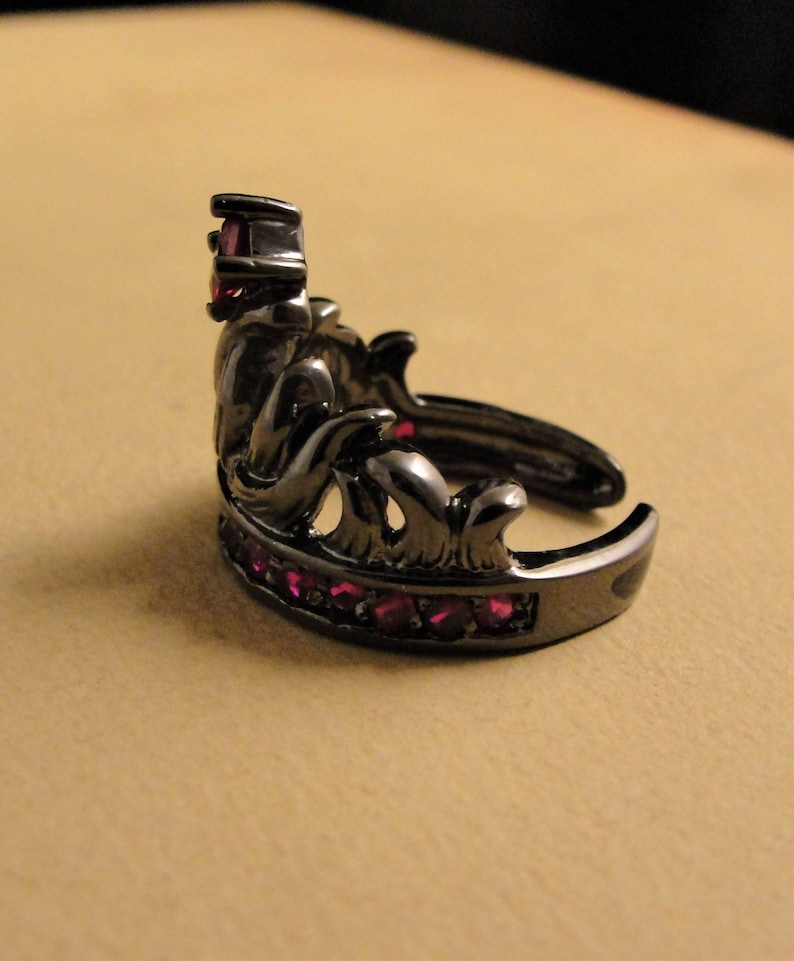 Peut inclure: Une bague couronne noire avec une seule pierre rouge sur le dessus et une rang&eacute;e de petites pierres rouges autour de la bande.