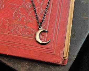 May include: A silver crescent moon pendant necklace with a dark chain. The necklace rests on a vintage red book with gold-edged pages. The background includes a dark, weathered book.
