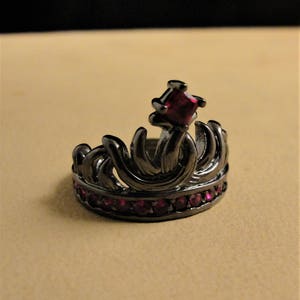 Peut inclure: Une bague couronne noire avec une grosse pierre rouge et de plus petites pierres rouges autour de la bande.