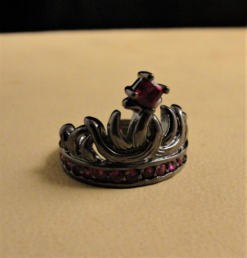 Peut inclure: Une bague couronne noire avec une grosse pierre rouge et de plus petites pierres rouges autour de la bande.
