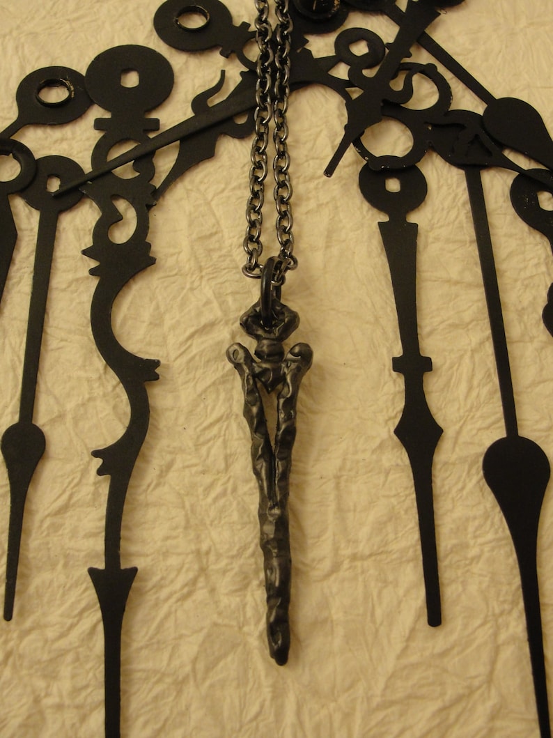 May include: A black metal pendant necklace with a long, thin, textured design. The pendant is hanging from a silver chain.