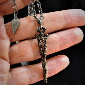 May include: A silver chain necklace with a detailed, textured, sword-shaped pendant. The pendant is about 2 inches long and has a pointed tip. The chain is about 20 inches long and has a lobster clasp.