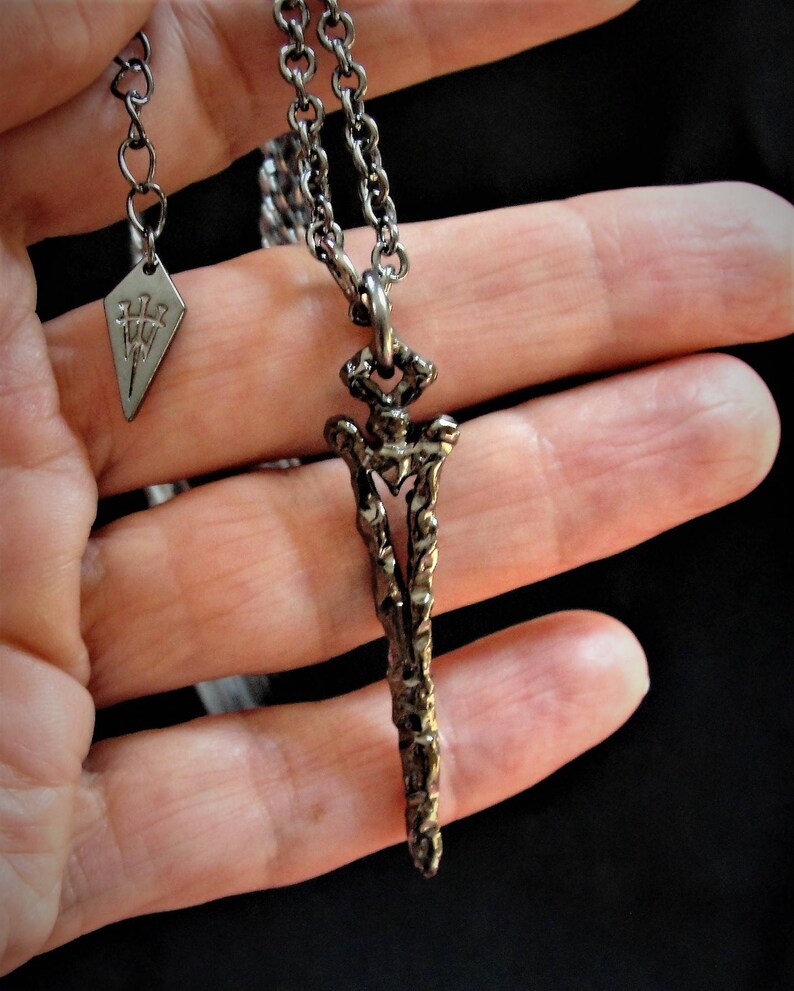 May include: A silver chain necklace with a detailed, textured, sword-shaped pendant. The pendant is about 2 inches long and has a pointed tip. The chain is about 20 inches long and has a lobster clasp.