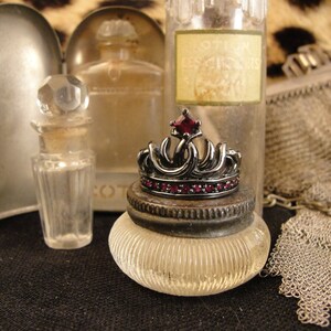Peut inclure: Une bague en forme de couronne noire avec une grande pierre rouge au centre. La bague est pos&eacute;e sur un pot en verre transparent &agrave; surface textur&eacute;e. Le pot est entour&eacute; d'autres bouteilles en verre et d'un sac en maille.