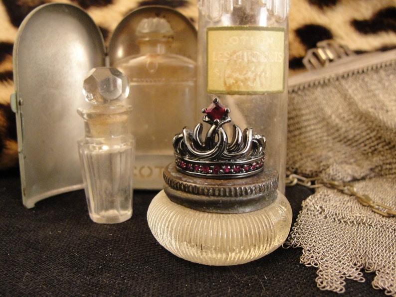 Peut inclure: Une bague en forme de couronne noire avec une grande pierre rouge au centre. La bague est pos&eacute;e sur un pot en verre transparent &agrave; surface textur&eacute;e. Le pot est entour&eacute; d'autres bouteilles en verre et d'un sac en maille.