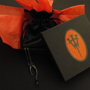 May include: A black box with a red oval logo containing a stylized letter "W" with three points. The box is open and a black velvet bag is inside. A silver necklace with a red gemstone pendant is hanging out of the bag. The box is sitting on a red tissue paper.
