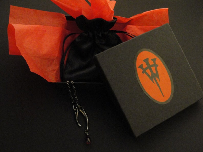 May include: A black box with a red oval logo containing a stylized letter "W" with three points. The box is open and a black velvet bag is inside. A silver necklace with a red gemstone pendant is hanging out of the bag. The box is sitting on a red tissue paper.
