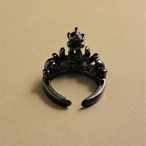 Peut inclure: Bague noire avec un motif de couronne et des pierres pr&eacute;cieuses roses. La bague est ouverte &agrave; l'arri&egrave;re et a un sommet pointu.