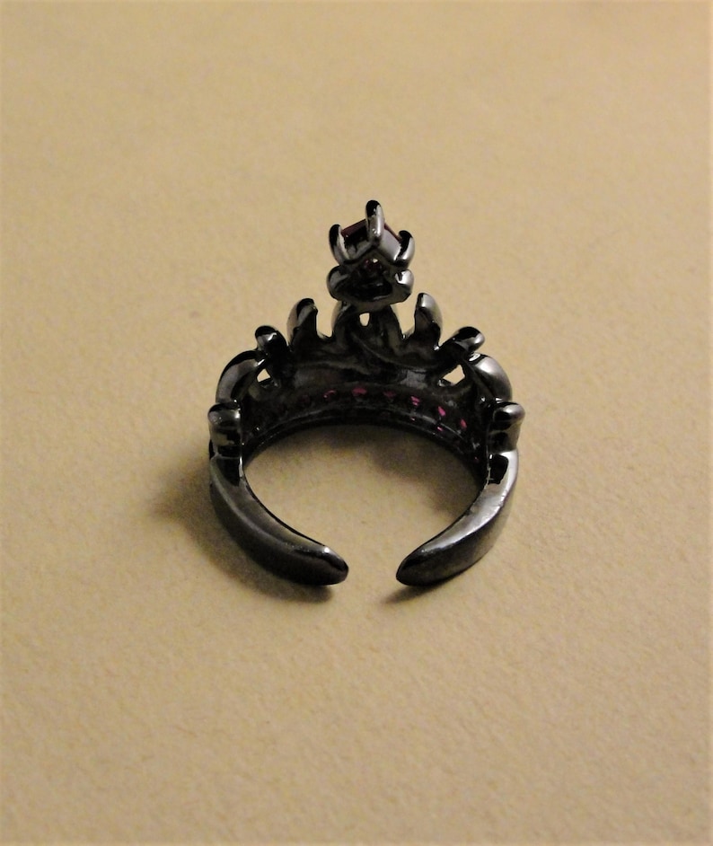 Peut inclure: Bague noire avec un motif de couronne et des pierres pr&eacute;cieuses roses. La bague est ouverte &agrave; l'arri&egrave;re et a un sommet pointu.