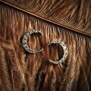 Op de afbeelding: Een paar zilveren halve maan oorbellen met heldere kristallen accenten op een bruine veren achtergrond.