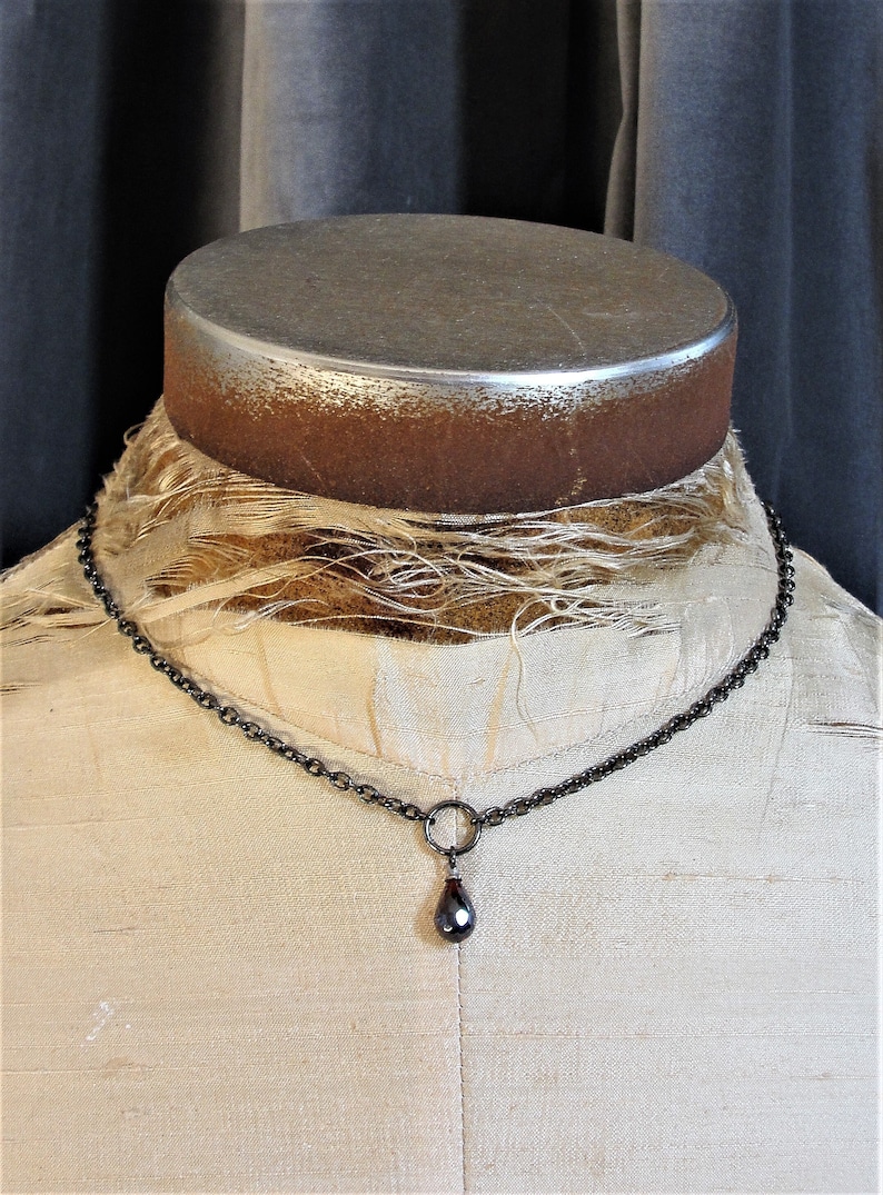 May include: A black chain necklace with a teardrop-shaped pendant. The pendant is a dark red color with a faceted surface.
