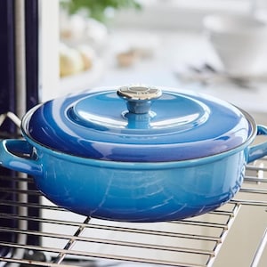 May include: A blue enameled cast iron Dutch oven with a matching lid. The pot has two handles and sits on a metal rack inside an oven. The lid has a silver knob. The color gradient ranges from light to dark blue.