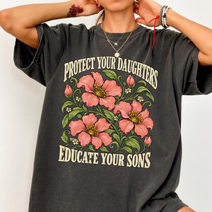 May include: A charcoal gray short-sleeve t-shirt featuring a graphic print of pink flowers with green leaves. The text "PROTECT YOUR DAUGHTERS" is printed in a cream-colored arched font above the flowers, and "EDUCATE YOUR SONS" is below.