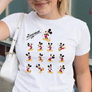 Może przedstawiać: Biała koszulka z grafiką Myszki Miki. Projekt zawiera wiele obraz&oacute;w Myszki Miki w r&oacute;żnych pozach, z napisem "Mickey Mouse" powyżej. Koszulka ma klasyczny okrągły dekolt.