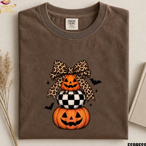 Op de afbeelding: Een bruin t-shirt met korte mouwen en een afbeelding van drie gestapelde items: een pompoen met gezicht, een zwart-wit geblokte bol en nog een pompoen, bekroond met een grote strik met luipaardprint en kleine zwarte silhouetten van vleermuizen.