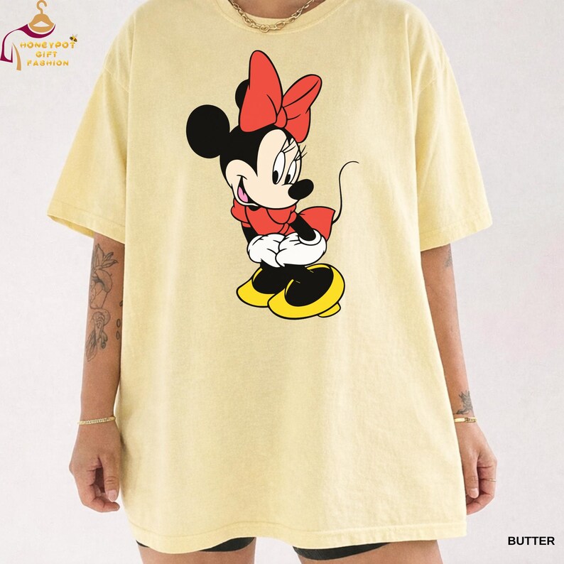 May include: Butter-colored t-shirt featuring a Minnie Mouse graphic. The cartoon character is depicted in her classic outfit: a red bow, red top, white gloves, and yellow shoes. The shirt has a relaxed fit and short sleeves. The text "HONEYPOT GIFT FASHION" is visible.