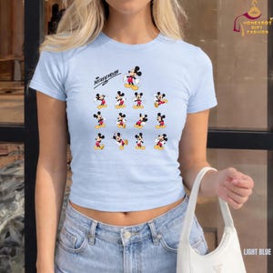 Może przedstawiać: Jasnoniebieski top z nadrukiem Myszki Miki. Koszulka ma okrągły dekolt i kr&oacute;tkie rękawy. Grafika Myszki Miki zawiera napis "Mickey Mouse" i wiele obraz&oacute;w Myszki Miki w r&oacute;żnych pozach.