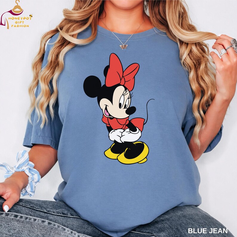 May include: A blue t-shirt featuring a Minnie Mouse graphic. Minnie is depicted in her classic outfit: a red bow, red dress, white gloves, and yellow shoes. The text "BLUE JEAN" is printed at the bottom right of the shirt.