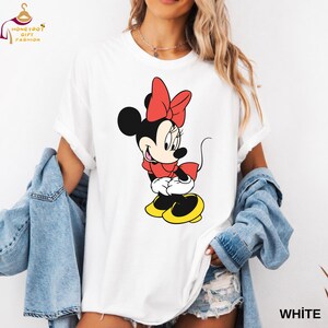 May include: White t-shirt featuring a colorful Minnie Mouse graphic. The cartoon character is depicted in a red dress and bow, with yellow shoes. The shirt has the word "WHITE" printed on the bottom right.