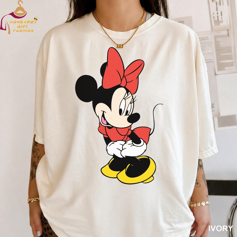 May include: Ivory-colored t-shirt featuring a Minnie Mouse graphic. The cartoon character is depicted in a red dress and bow, black ears, and yellow shoes. The shirt has short sleeves and a relaxed fit. The text "IVORY" is printed at the bottom right.